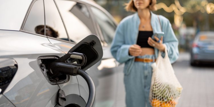 Cloae-up of fast charger plug in vehicle charging port, woman with groceries waiting for her car to be charged on background