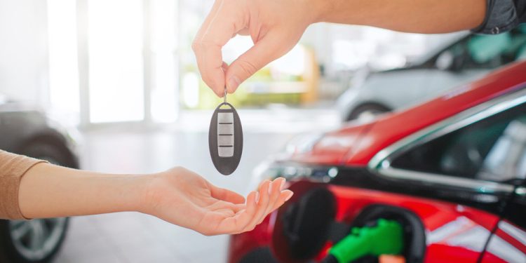 Young family buying first electric car in the showroom. Close-up of male hand giving car key to female hand on battery electric car background. Eco car sale in auto salon