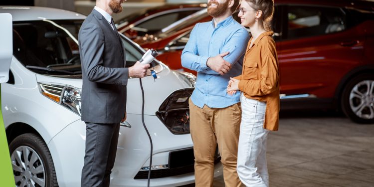 Sales manager showing car charging station to a young couple, selling electric cars in the showroom. Concept of buying eco-friendly car for family