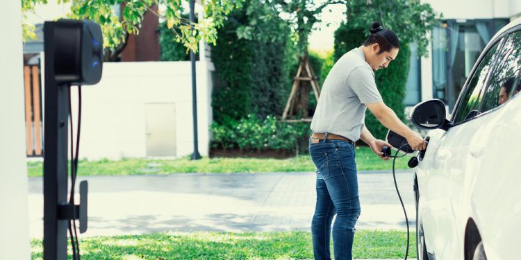 Progressive asian man install cable plug to his electric car with home charging station in the backyard. Concept use of electric vehicles in a progressive lifestyle contributes to clean environment.